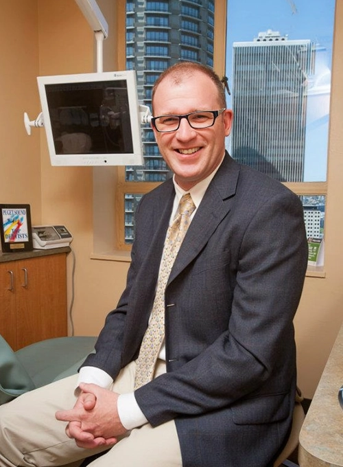 Dentist Thomas McKenny in the office smiling in suit and yellow tie with skyscrapers in background